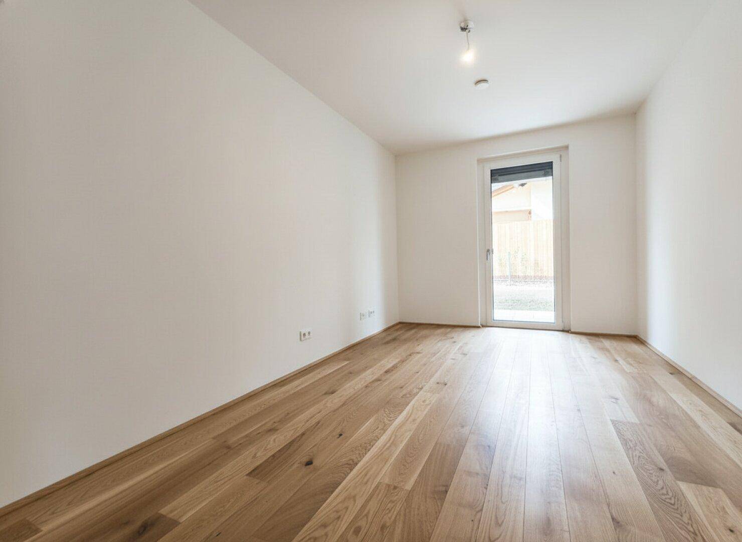 Leeres Zimmer mit Holzboden und Fenster, das viel Tageslicht hereinlässt.