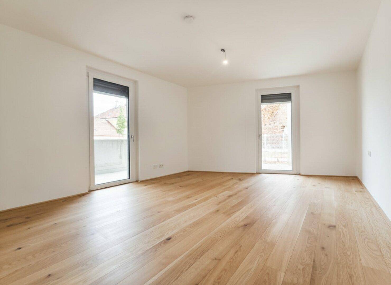 Geräumiges Zimmer mit Holzboden und zwei Fenstern, die auf einen Balkon führen.