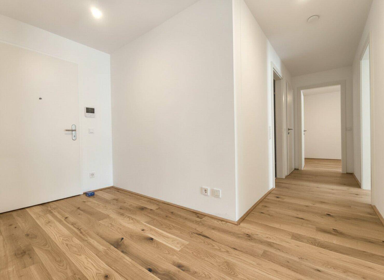 Heller Eingangsbereich mit Holzdielenboden und weißer Eingangstür, die zu weiteren Räumen führt.