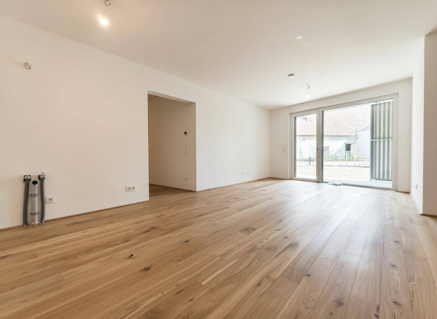 Heller, leerer Wohnraum mit breiten Holzdielen und Zugang zu einem Außenbereich durch eine Glasschiebetür.