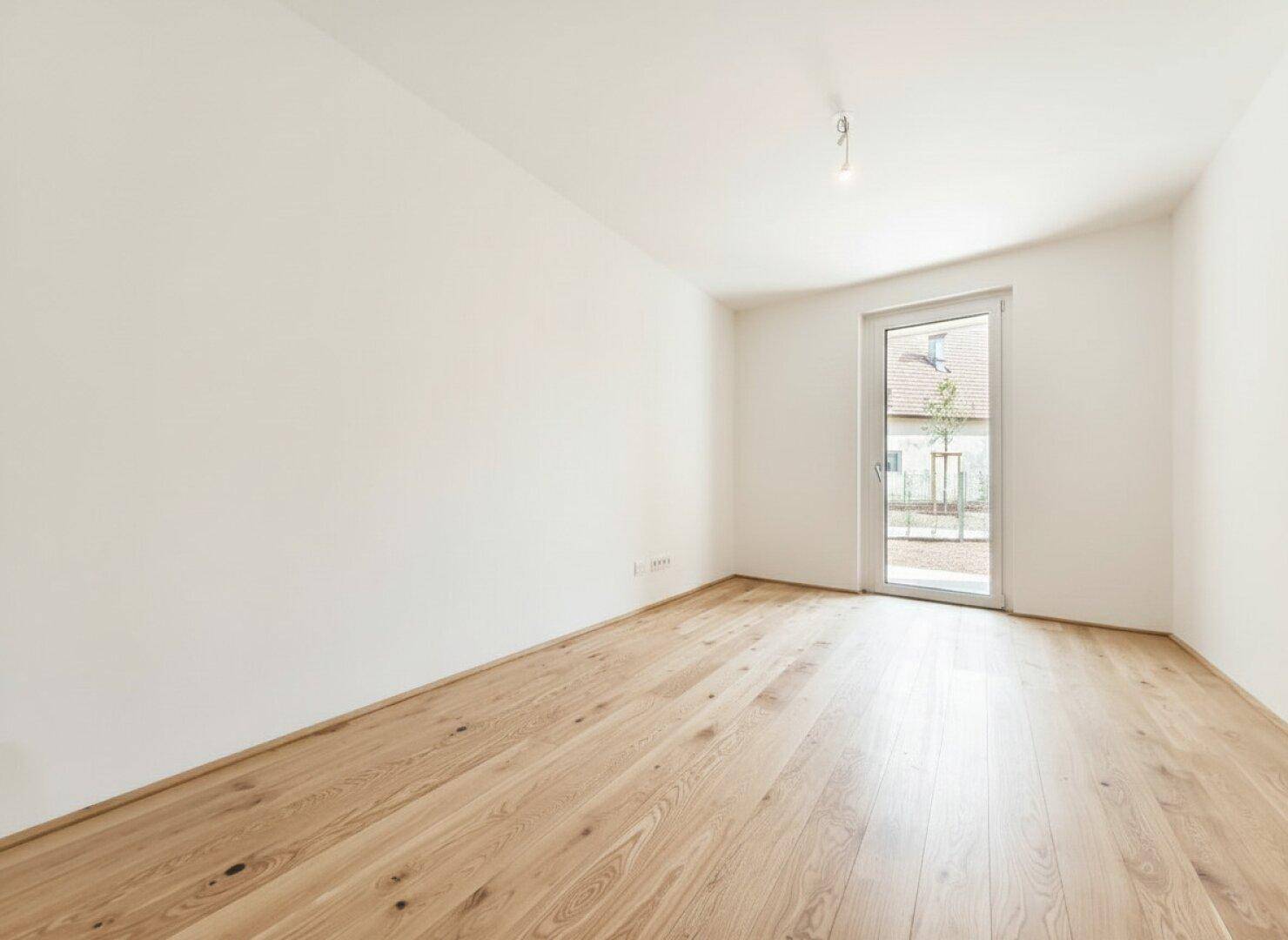 Unmöblierter Raum mit warmen Holzdielen und einer Glastür, die viel Tageslicht hereinlässt.