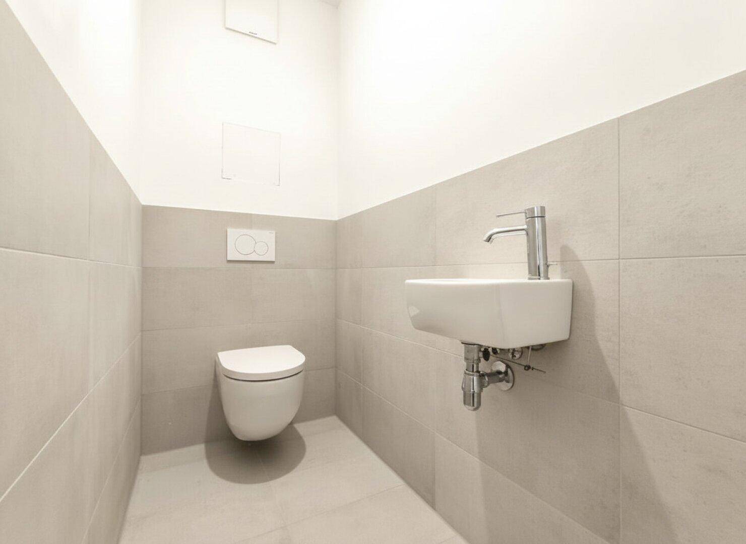 Gäste-WC mit modernen hellgrauen Fliesen, wandhängender Toilette und einem kleinen Handwaschbecken.