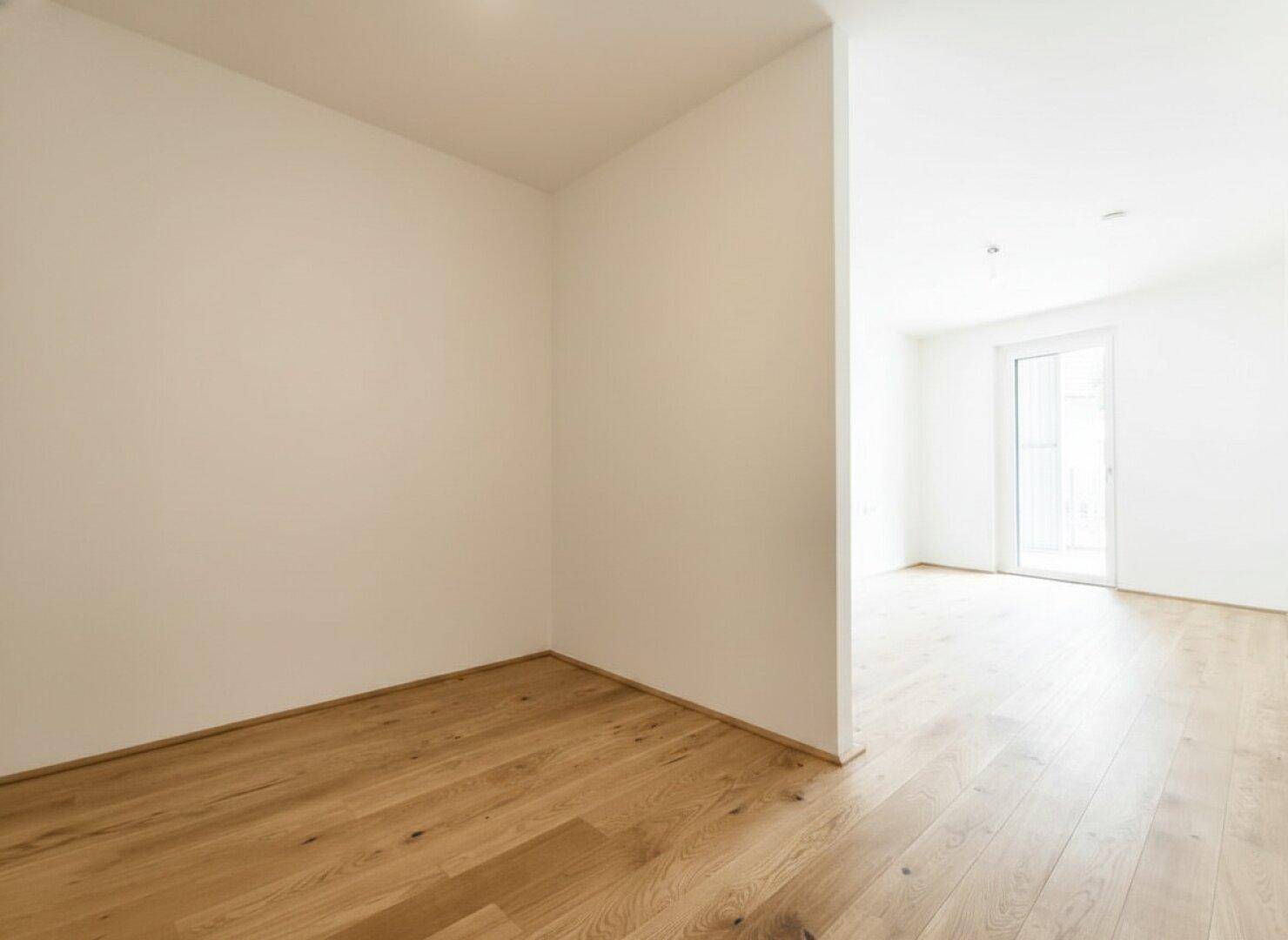 Helle Ecke eines unmöblierten Raumes mit Holzdielenboden und Übergang zu einem weiteren Bereich.