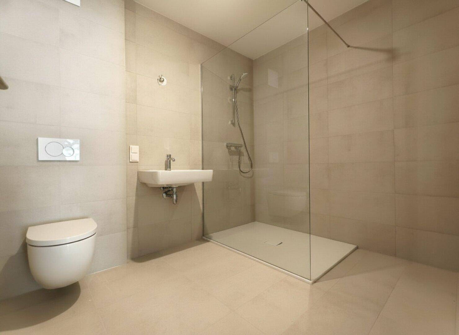 Modernes Badezimmer mit hellgrauen Fliesen, wandhängendem WC und einer geräumigen begehbaren Dusche.