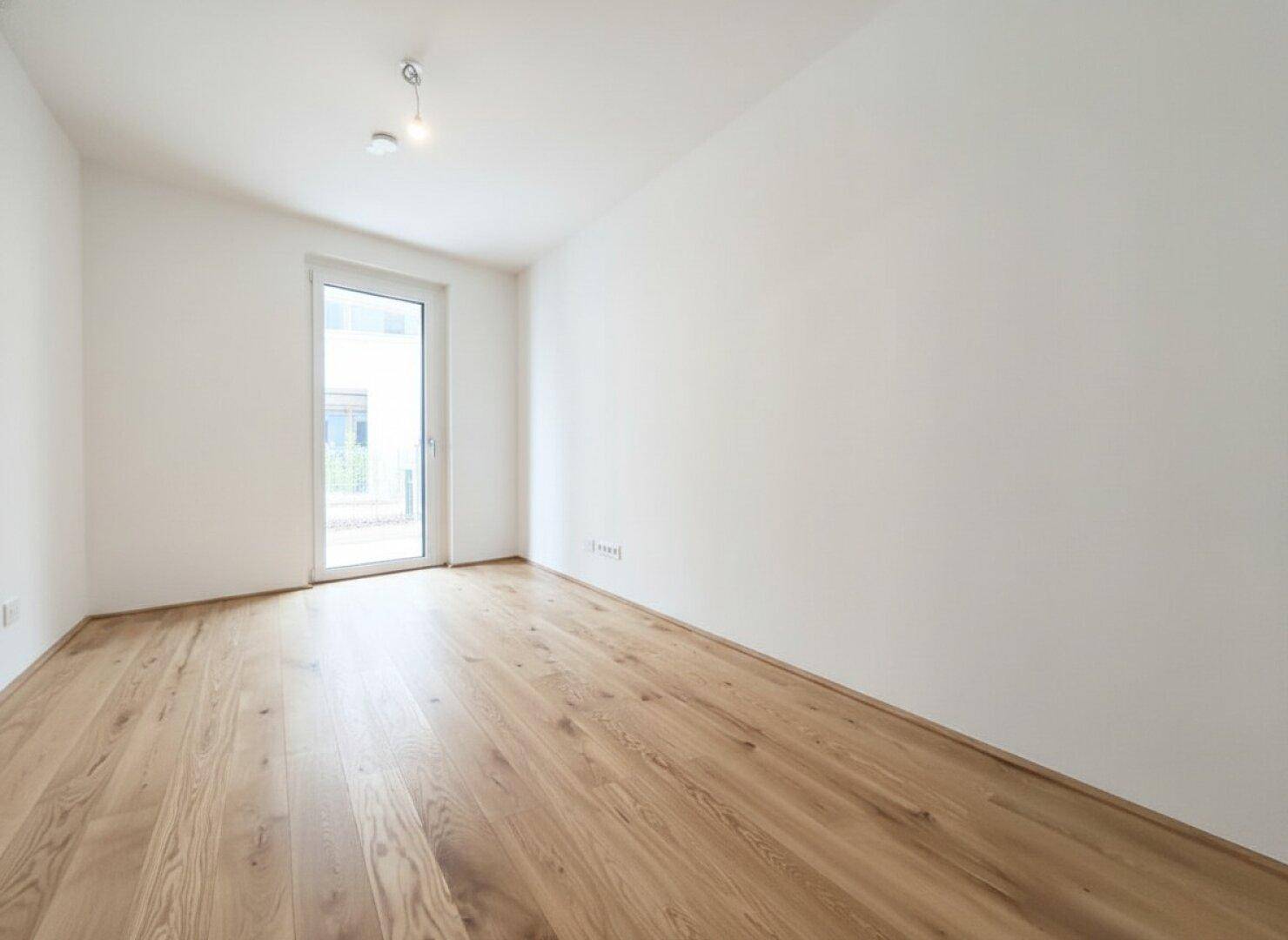 Geräumiges, leeres Zimmer mit Holzboden und Fenster-Tür-Kombination für viel Licht.