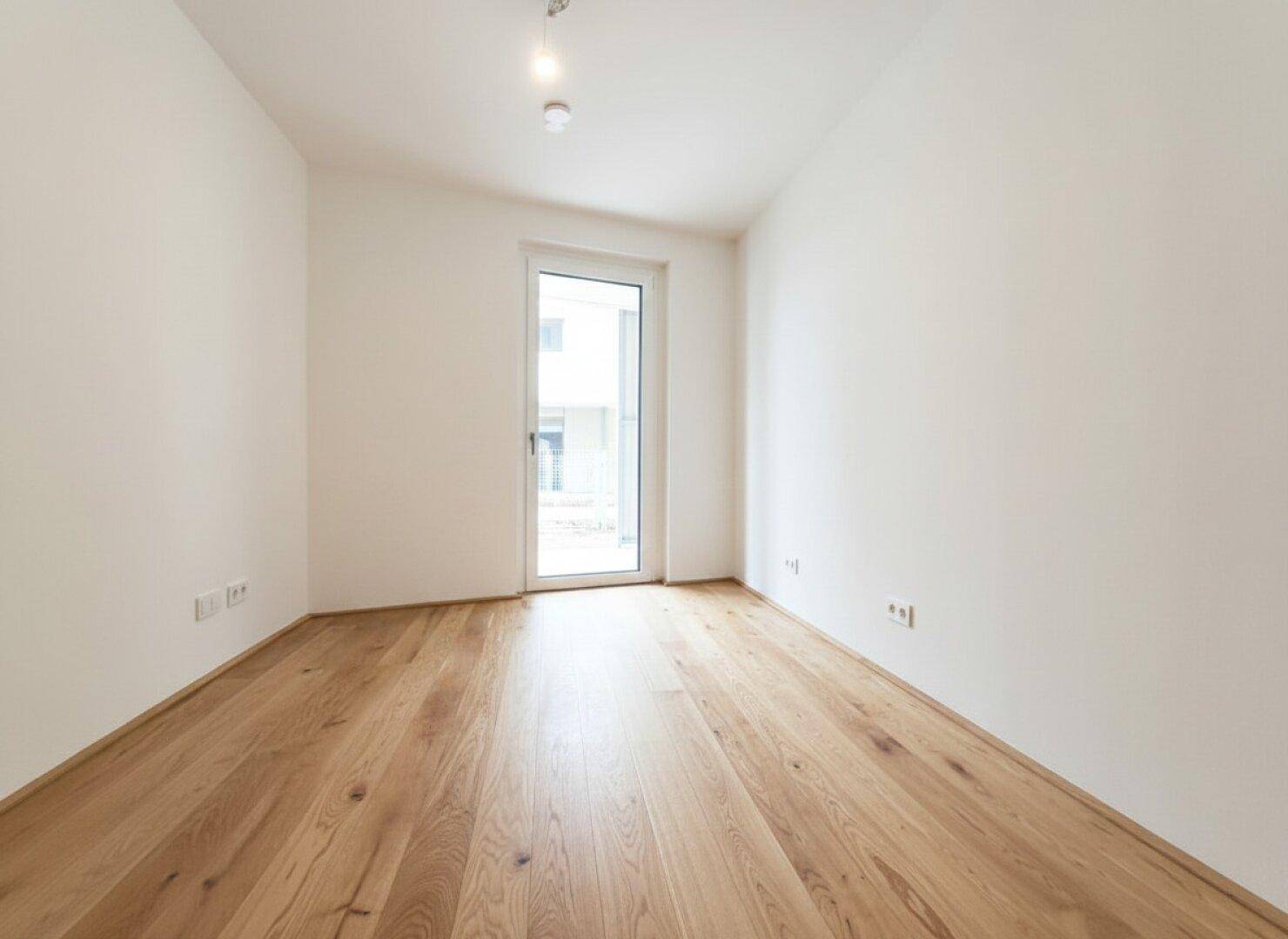 Unmöbliertes Zimmer mit warmem Holzboden und Zugang zu einem Balkon oder einer Loggia.