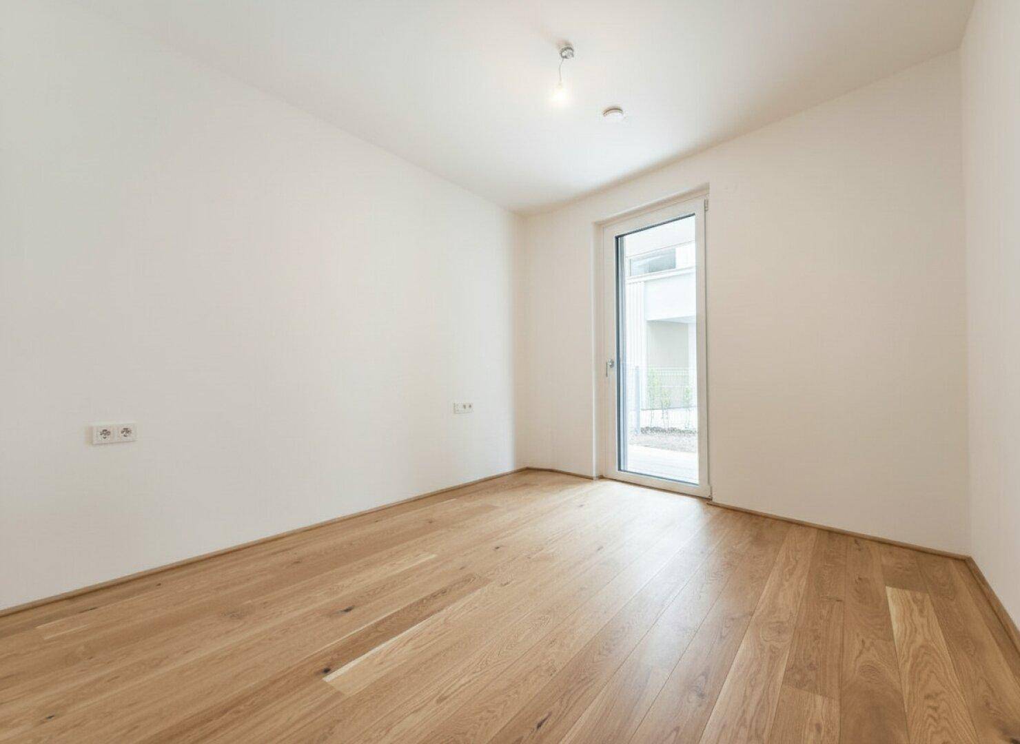 Unmöbliertes Schlafzimmer mit hellem Holzparkett und einer Tür, die zum Außenbereich führt.