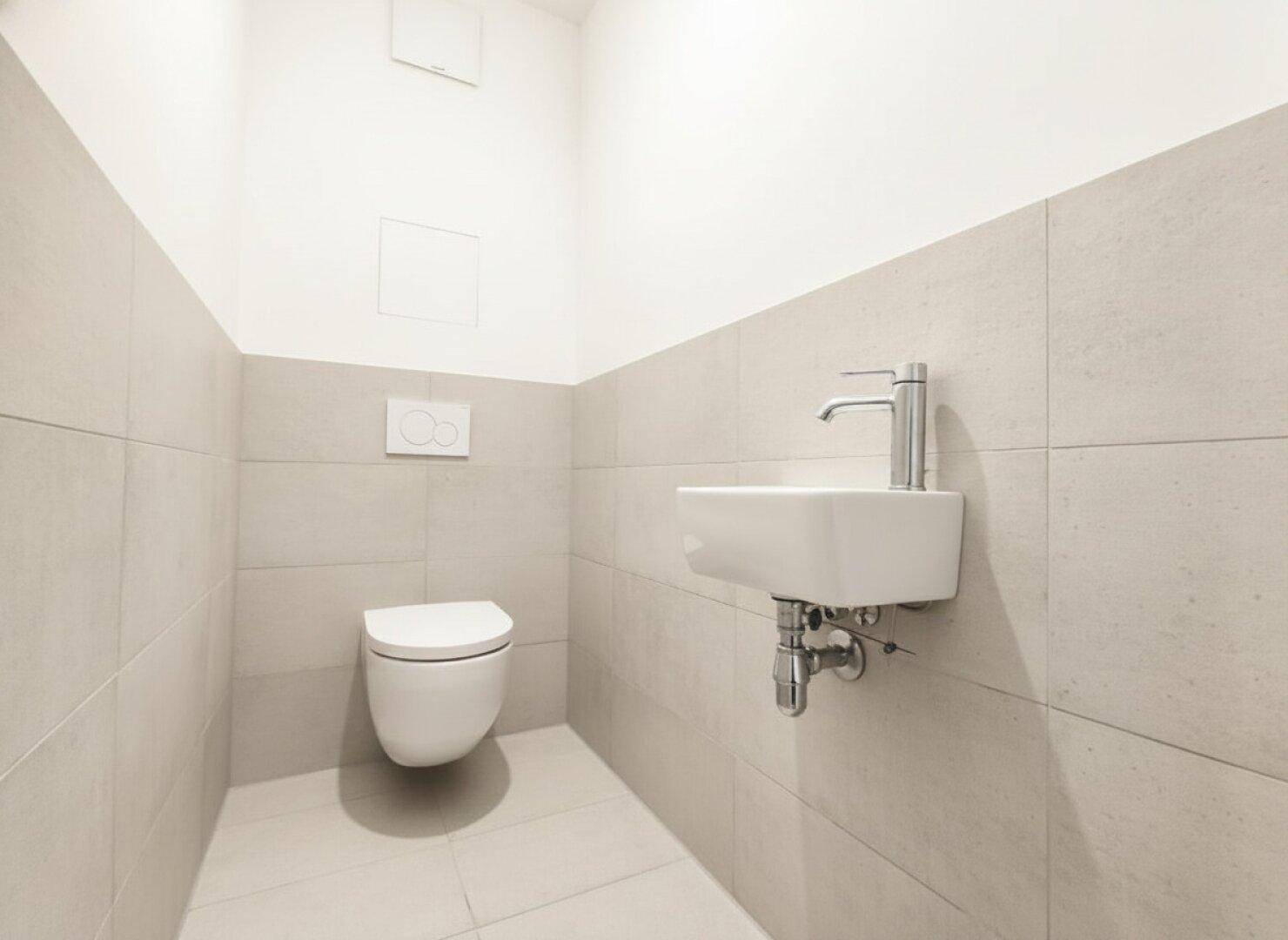 Modernes Gäste-WC mit grauen Fliesen, wandhängendem WC und kleinem Waschbecken.