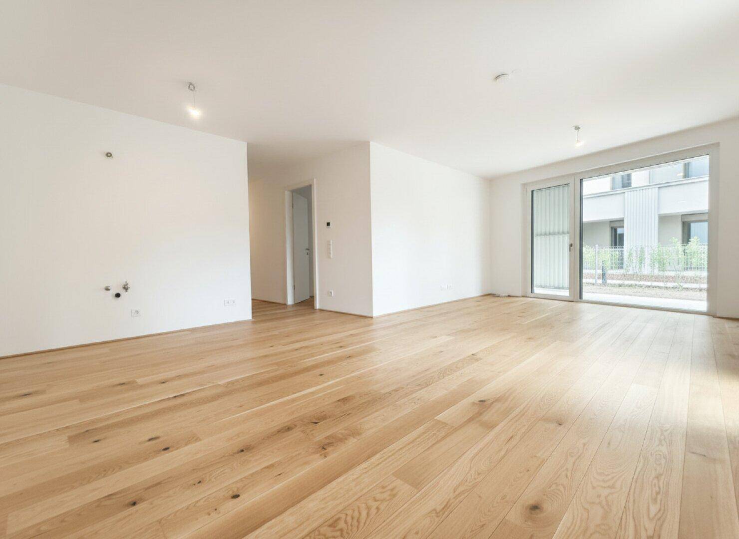 Heller, leerer Wohnraum mit hellem Holzfußboden und großer Schiebetür zum Außenbereich.