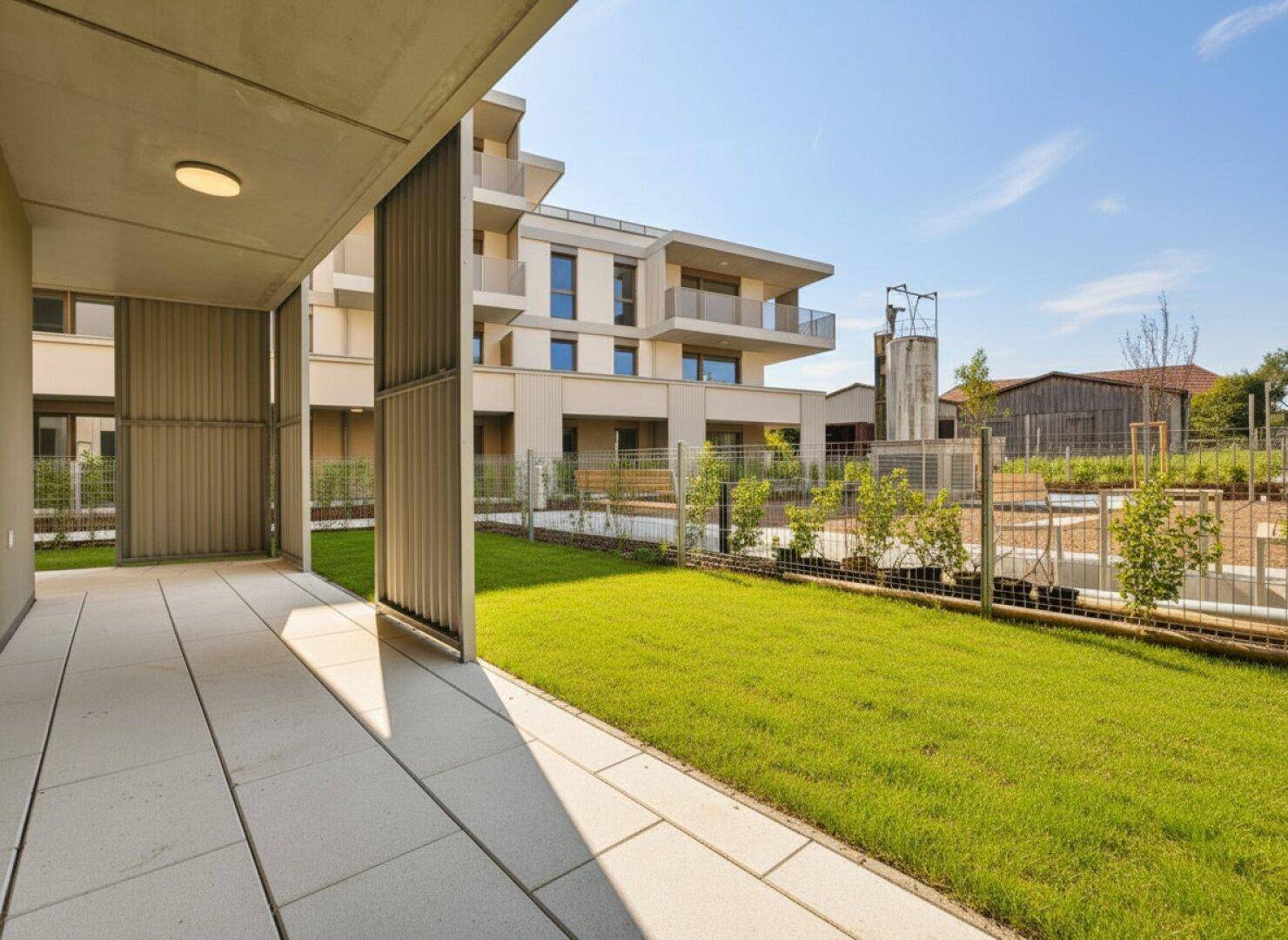 Gepflasterte Terrasse mit Rasenfläche und Blick auf ein modernes Wohngebäude unter blauem Himmel.