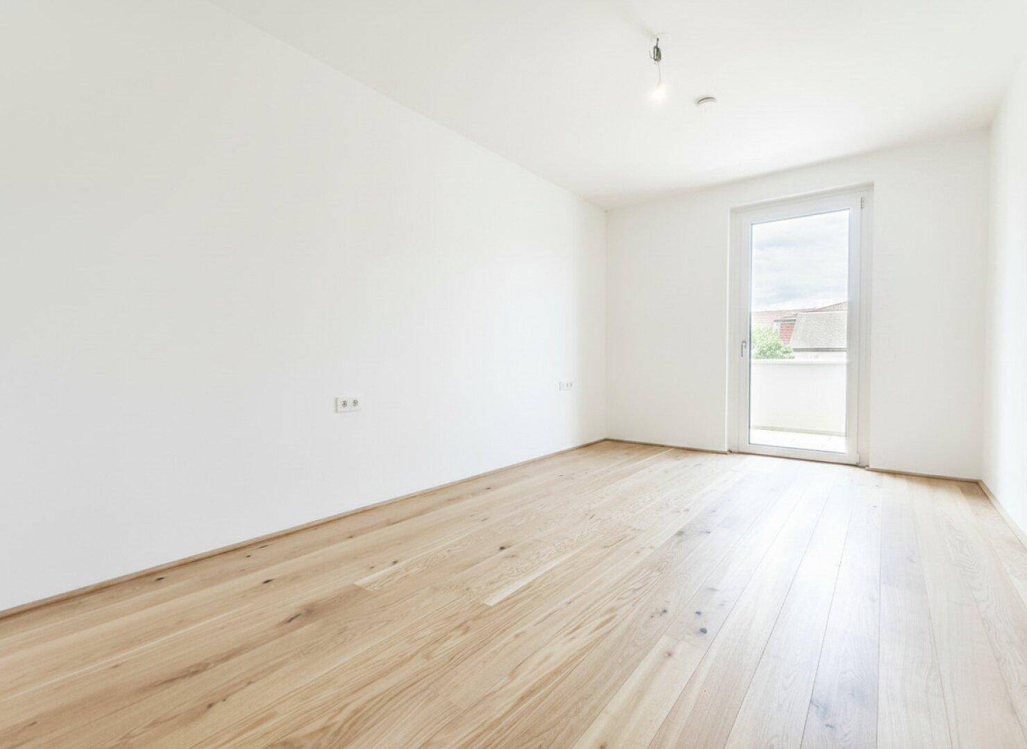 Leeres Zimmer mit hellem Holzboden und Fenster zum Balkon, bereit zur Einrichtung.