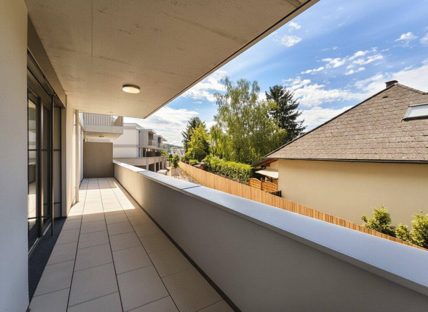 Langer, schmaler Balkon mit Fliesenboden und Blick auf Bäume und Nachbarhäuser.