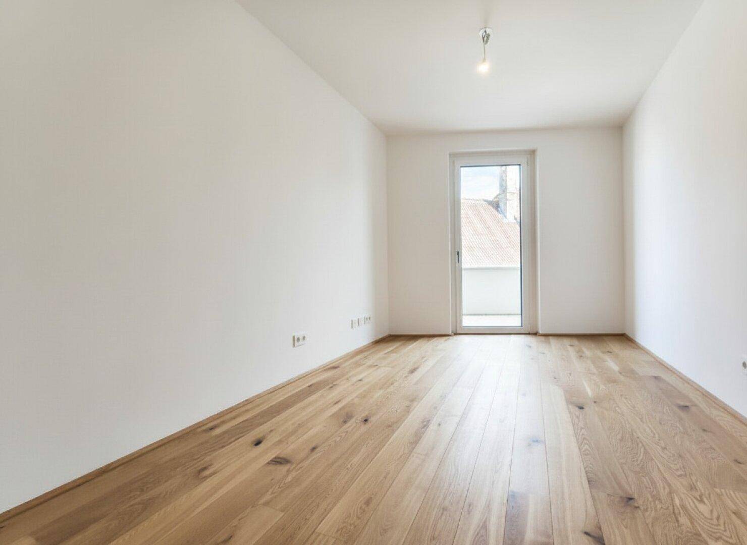 Geräumiges Schlafzimmer mit hellem Holzboden und viel Tageslicht durch die Balkontür.