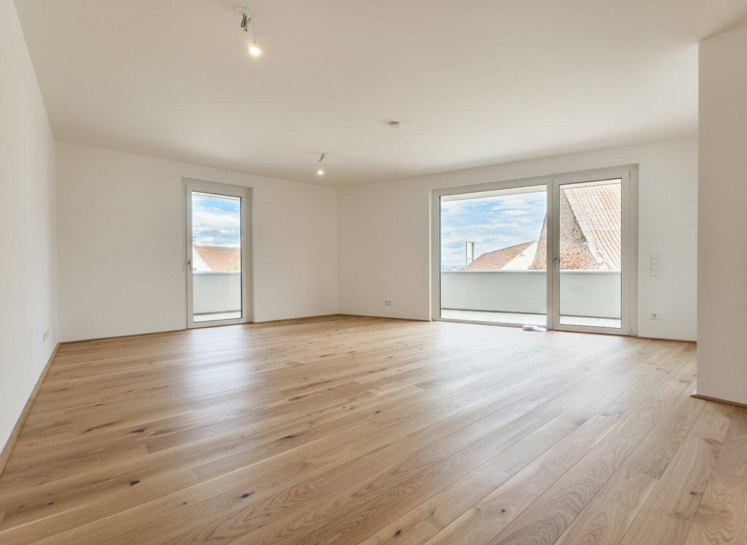 Geräumiger, heller Wohnbereich mit Holzboden und zwei großen Fenstern, die viel Tageslicht hereinlassen.