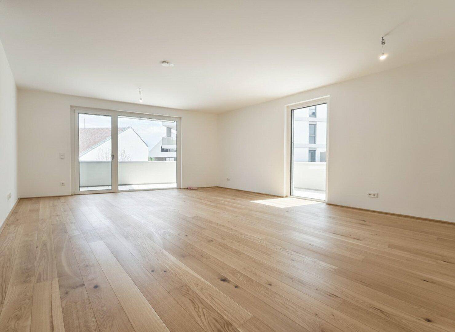 Helles, leeres Wohnzimmer mit Eichenparkett und Zugang zu einem Balkon.