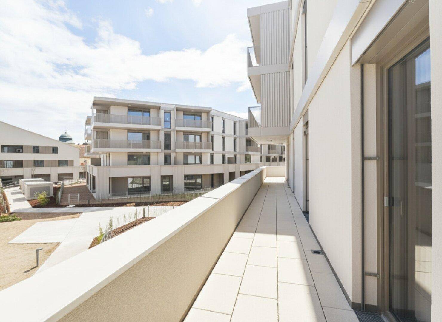Langer Balkon mit Fliesenboden und Blick auf moderne Wohngebäude unter blauem Himmel.