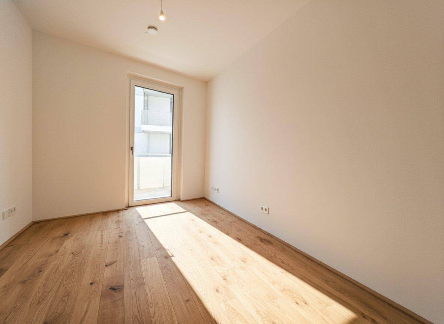 Sonniger Raum mit Holzboden und Balkonzugang, der viel natürliches Licht hereinlässt.