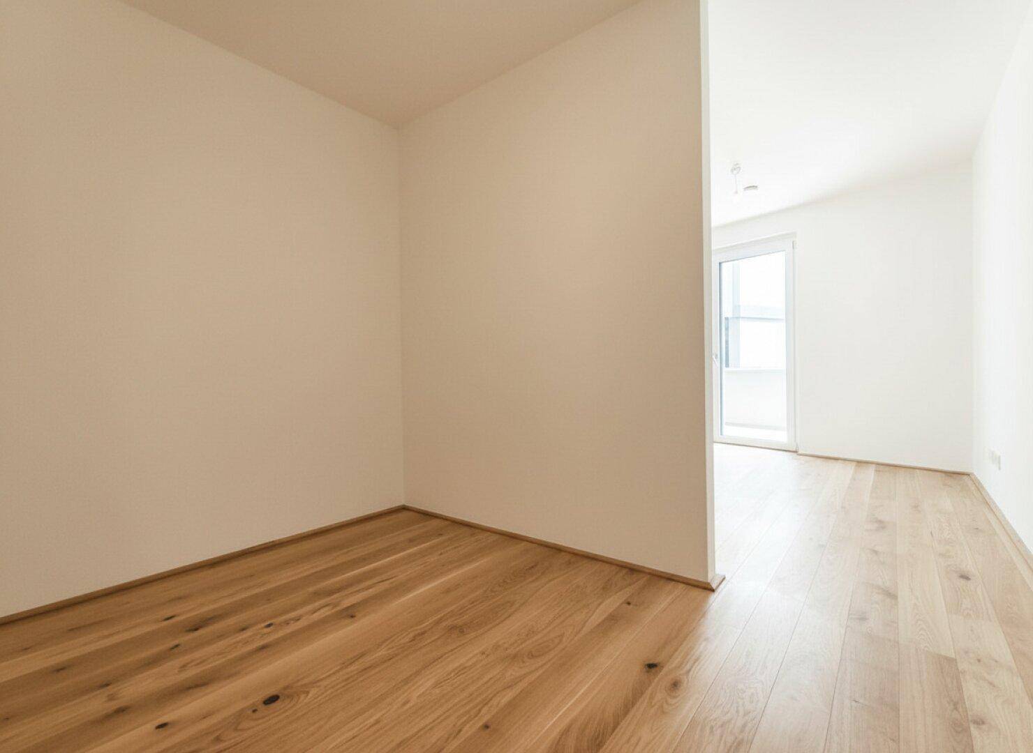 Heller, leerer Raum mit Holzboden, der vielseitig nutzbar ist und Zugang zu einem weiteren Bereich bietet.