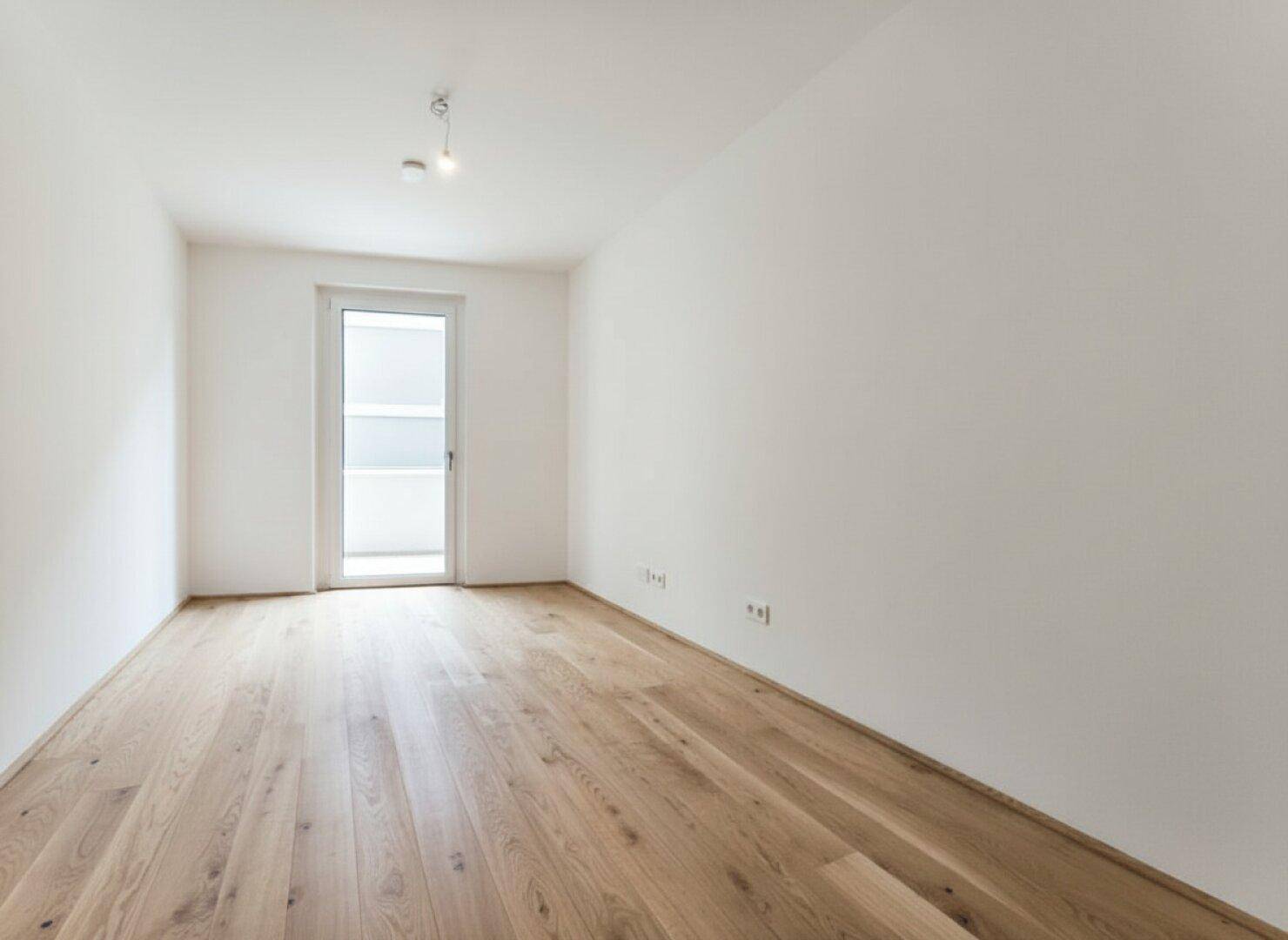 Leeres Zimmer mit Holzboden und Balkontür, bietet viel natürliches Licht und frische Luft.