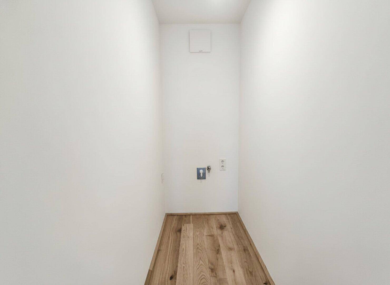 Schmaler Raum mit Holzboden und Anschlüssen für Waschmaschine, ideal als Hauswirtschaftsraum.