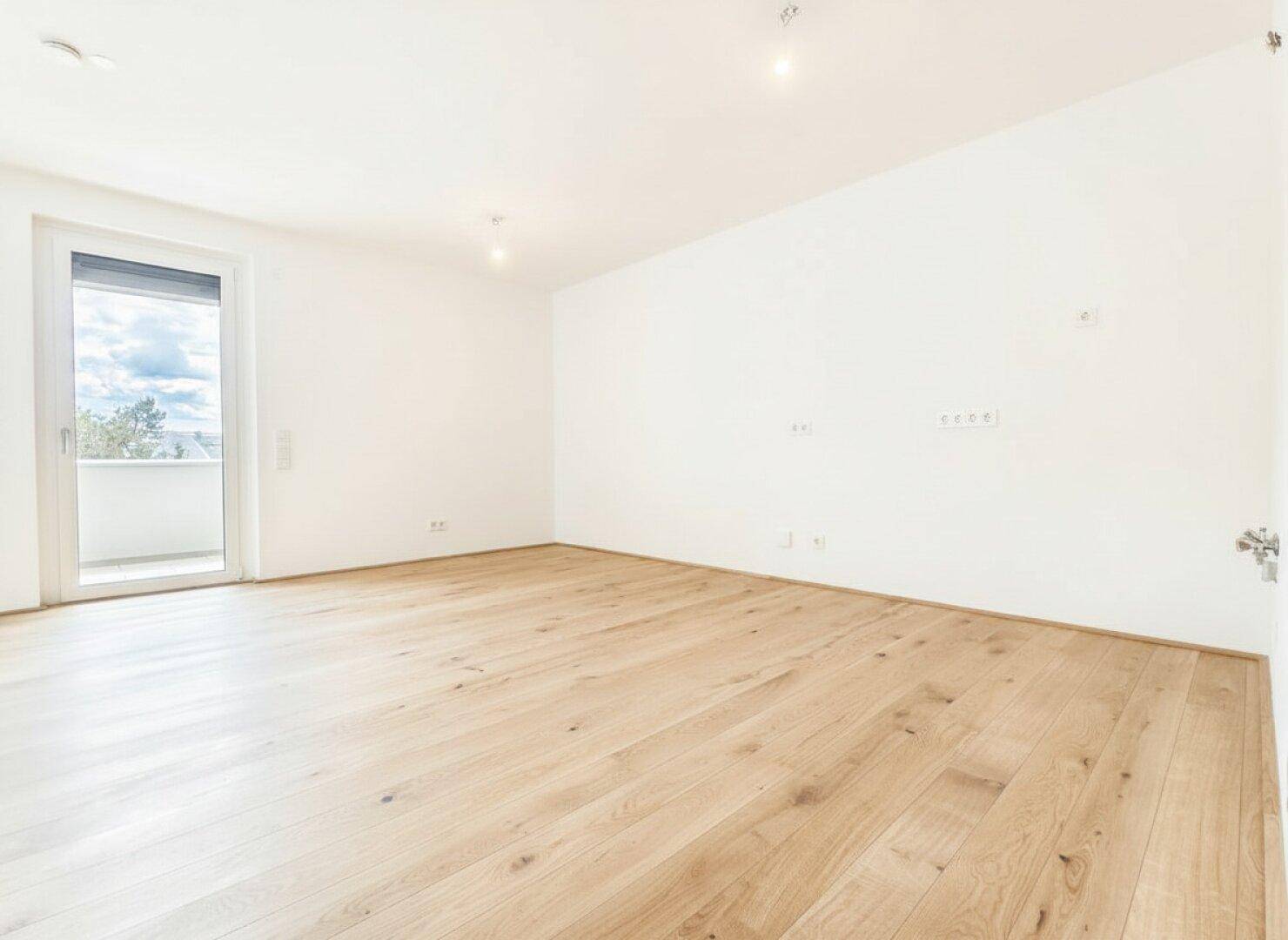 Lichtdurchfluteter Raum mit Holzboden und Zugang zu einem Balkon, bietet einen weiten Blick.