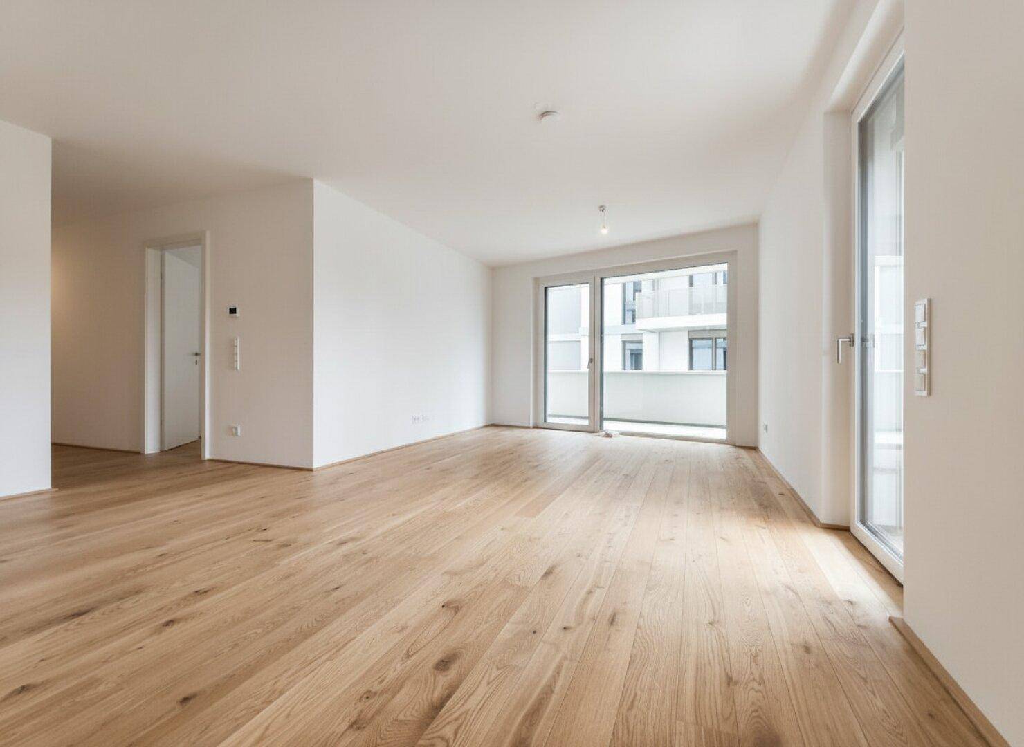 Helles, leeres Zimmer mit Holzboden und Zugang zu einem Balkon, ideal als Wohnbereich.