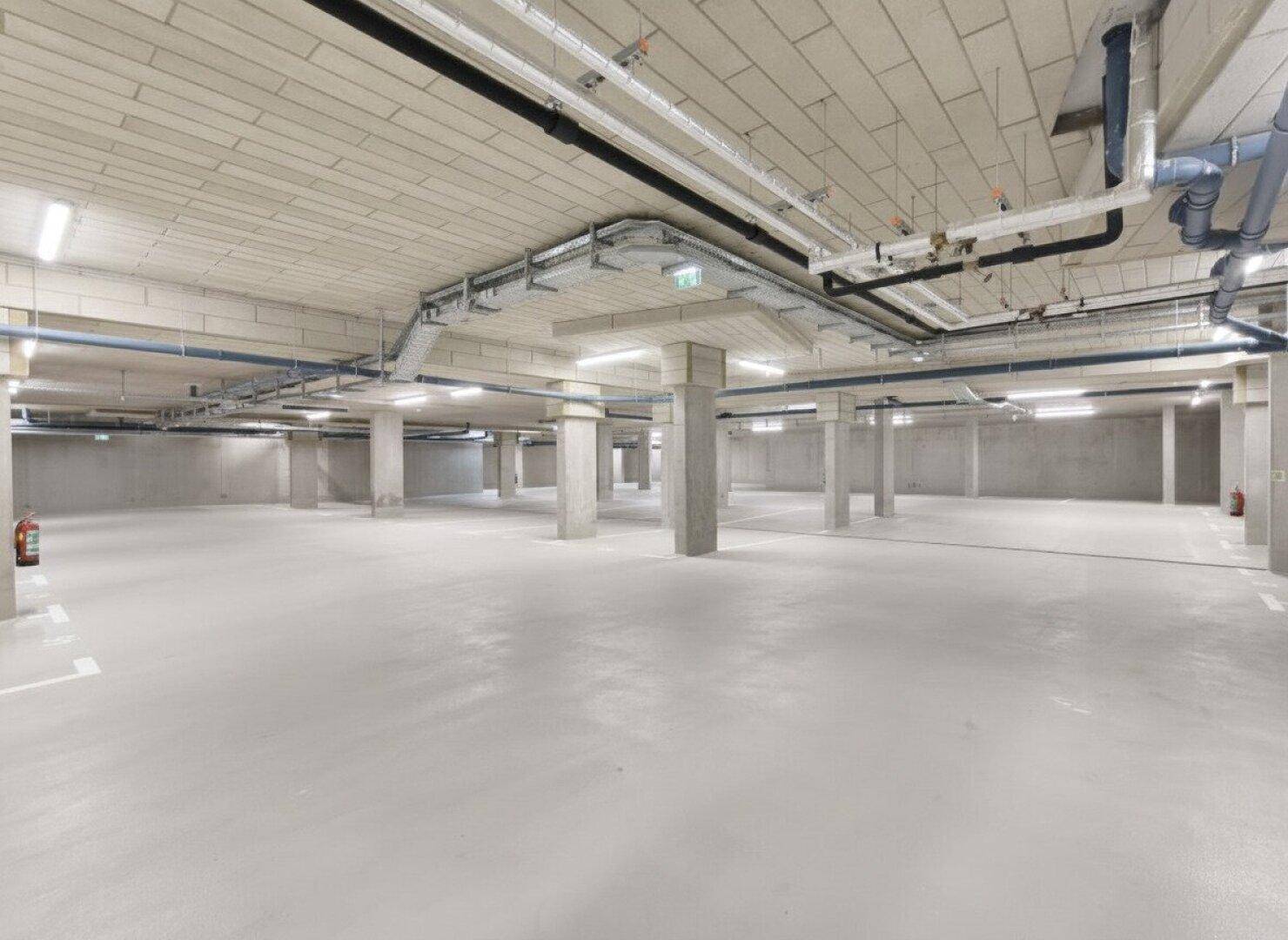 Große Tiefgarage mit vielen Stellplätzen, Betonpfeilern und sichtbaren Leitungen an der Decke.