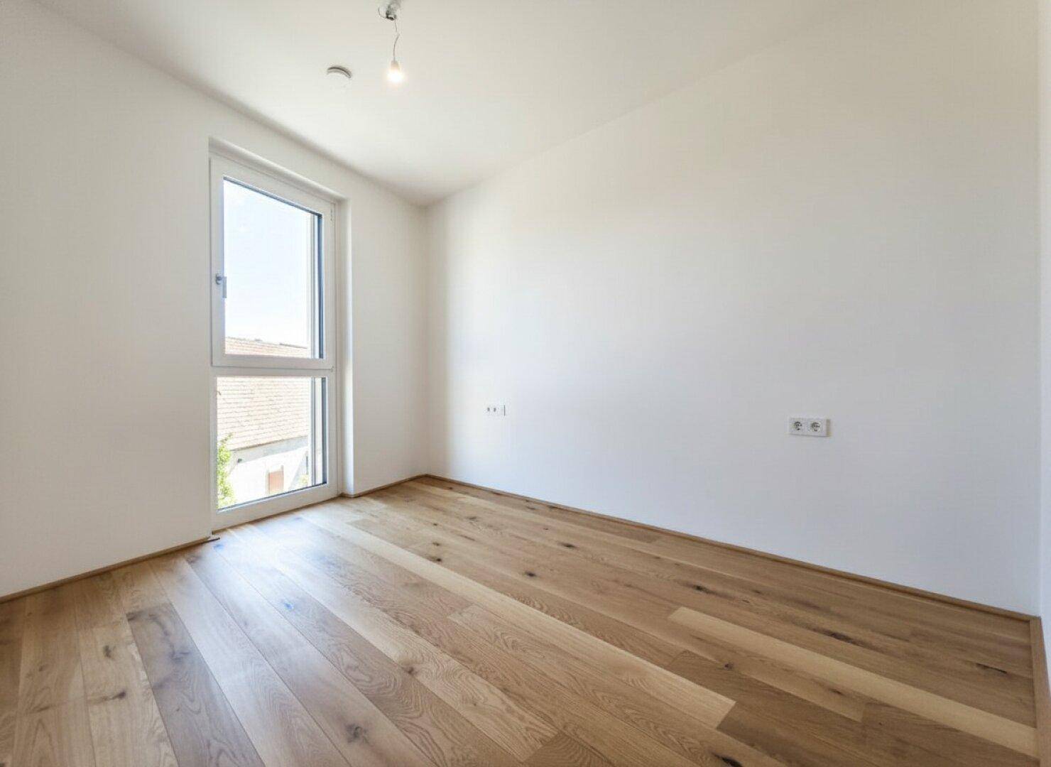 Leeres Schlafzimmer mit hellem Eichenparkett und einem Fenster mit Blick nach draußen.