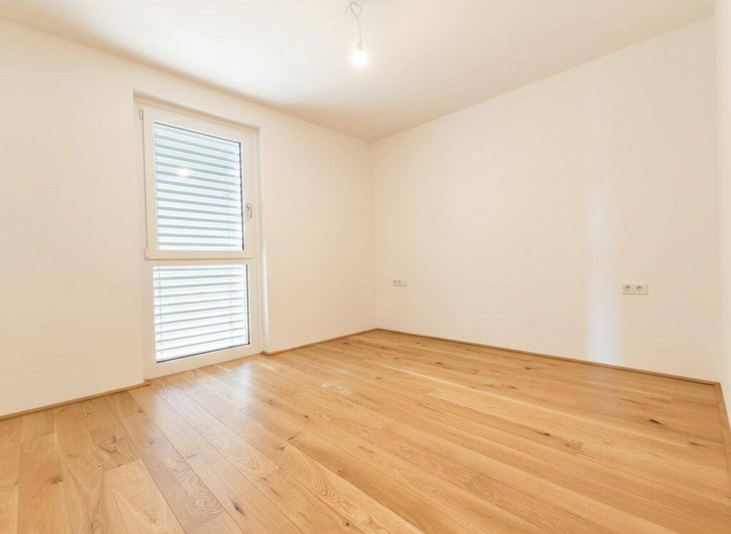 Helles Zimmer mit Parkettboden und Fenster mit Jalousien für Privatsphäre.