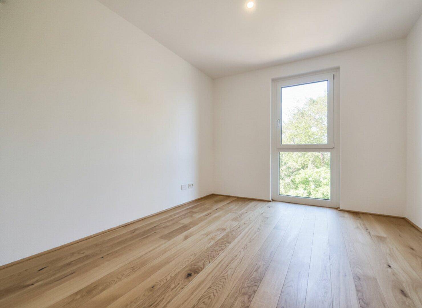 Helles Zimmer mit Holzboden und großem Fenster, bietet viel Tageslicht und eine angenehme Atmosphäre.