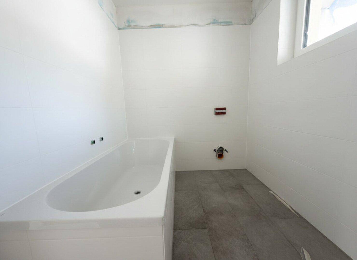 Unfertiges Badezimmer mit weißen Wandfliesen, grauen Bodenfliesen und einer eingebauten Badewanne.