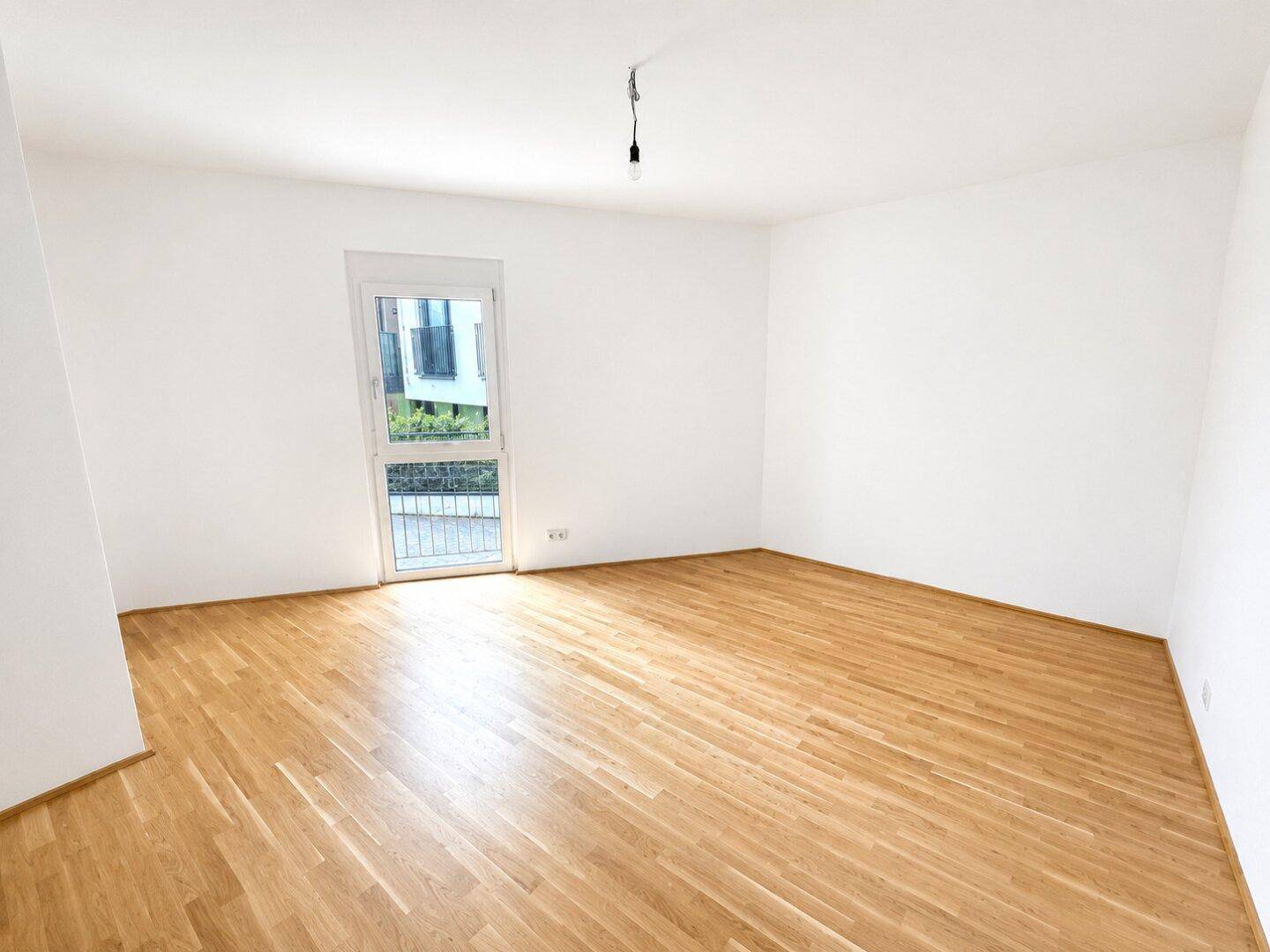 Leeres Zimmer mit Parkettboden und einem großen Fenster, das viel Tageslicht hereinlässt.