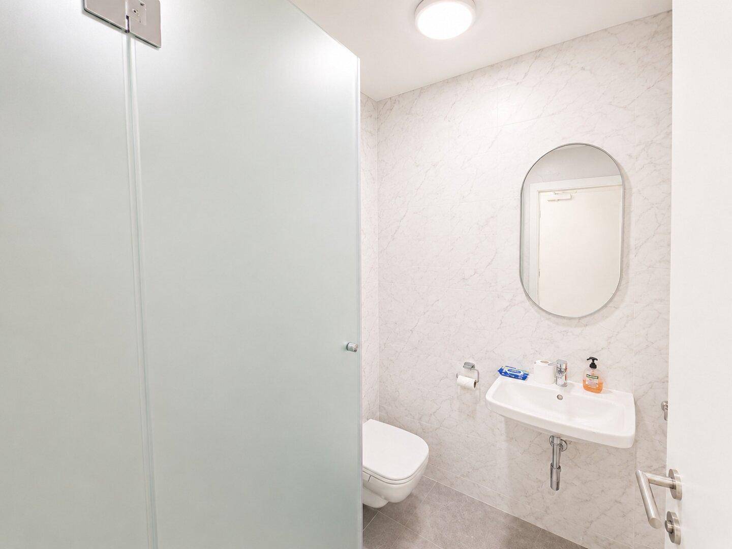 Modernes Badezimmer mit weißer Wandverkleidung, Hänge-WC und ovalem Spiegel über dem Waschbecken.