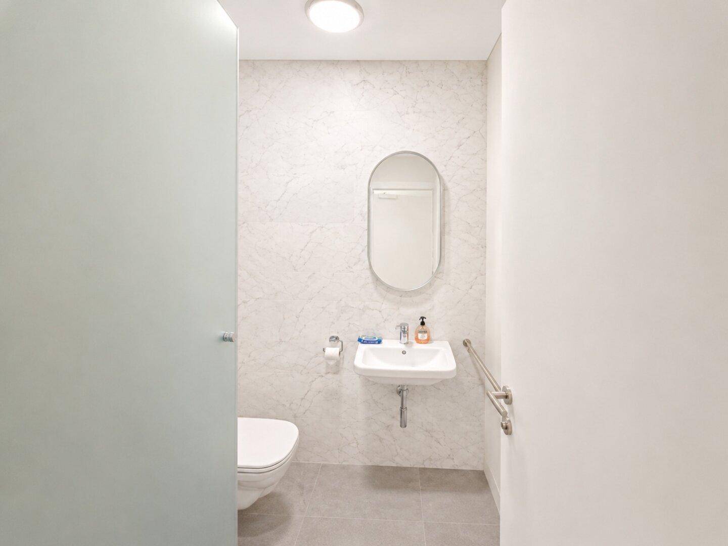 Barrierefreies Badezimmer mit modernen Fliesen, Hänge-WC, Waschbecken und Haltegriff an der Wand.