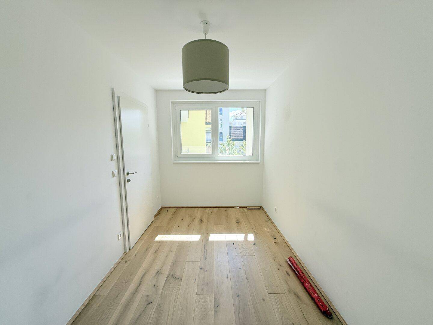 Leeres Schlafzimmer mit Holzboden, weiß gestrichenen Wänden und einem großen Fenster.