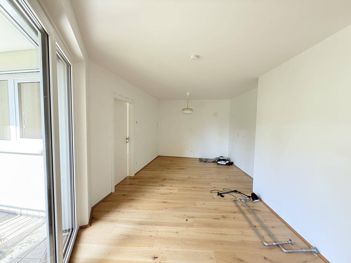 Heller, leerer Raum mit Parkettboden und großen Fenstern, die zum Balkon führen.