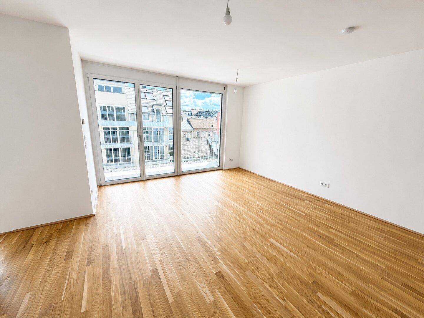 Helles, leeres Wohnzimmer mit Eichenparkettboden und großen Schiebetüren zum Balkon.