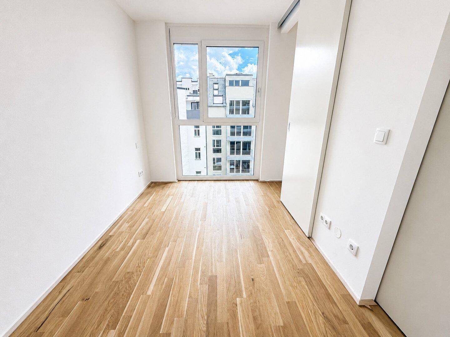 Helles Schlafzimmer mit Parkettboden und großem Fenster, das viel Tageslicht hereinlässt.