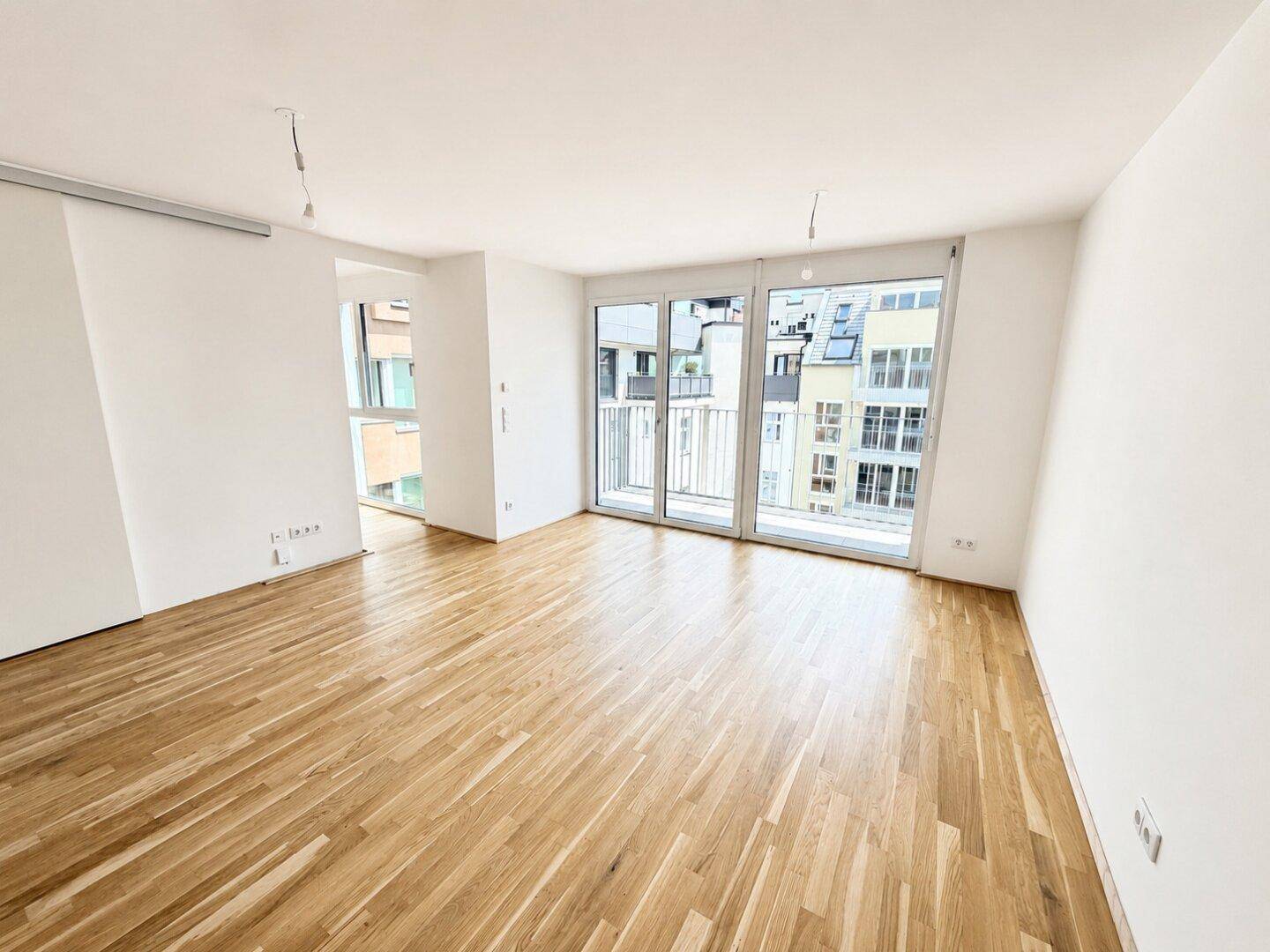 Geräumiges, helles Zimmer mit Parkettboden und Zugang zu einem Balkon, umgeben von modernen Gebäuden.