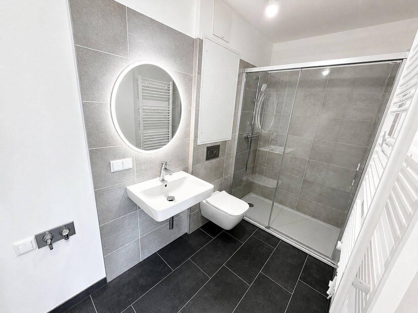 Modernes Badezimmer mit grauen Fliesen, begehbarer Dusche, wandhängendem WC und beleuchtetem Spiegel.