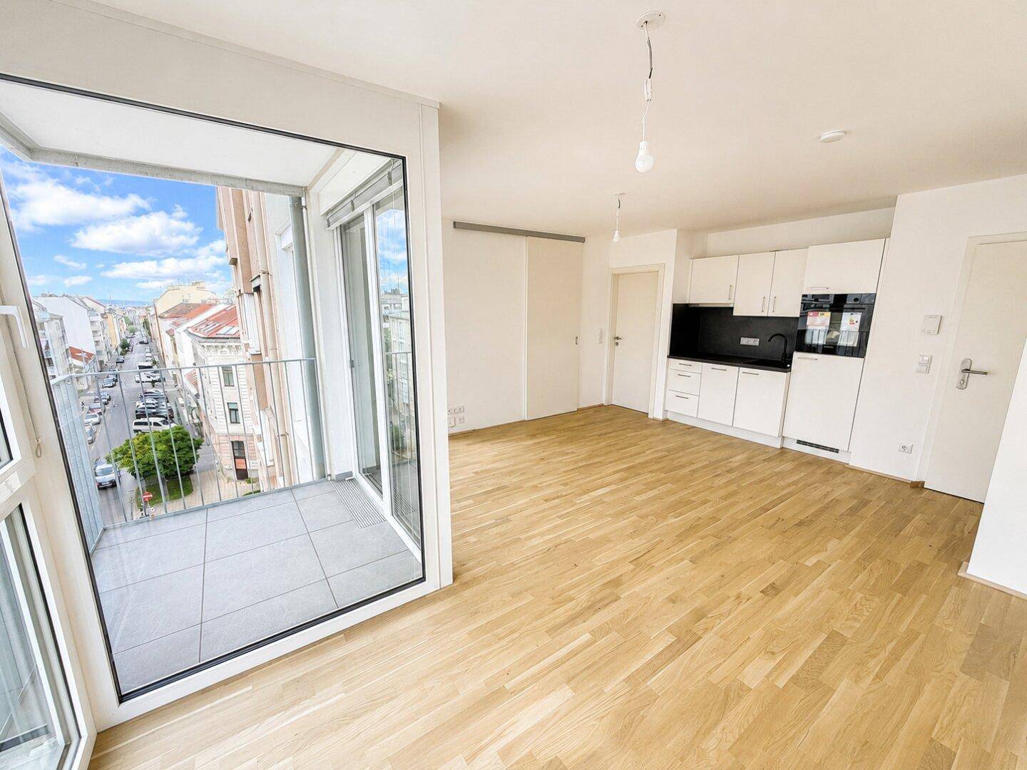 Offener Wohnbereich mit integrierter Küchenzeile, Parkettboden und Zugang zu einem gefliesten Balkon.