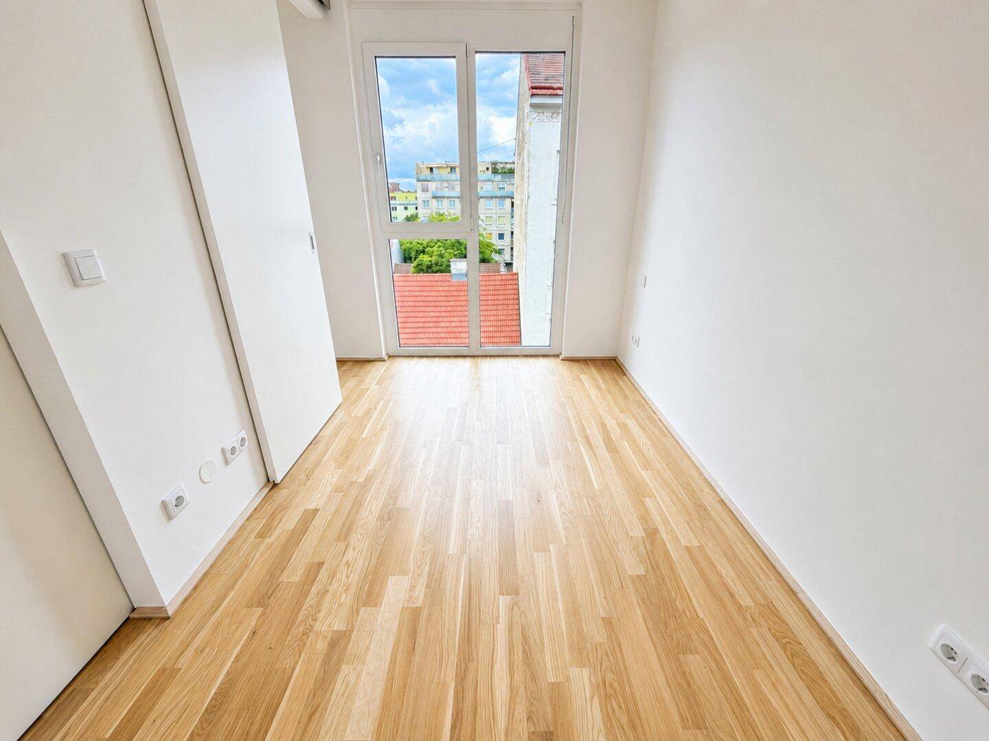 Ruhiges Schlafzimmer mit hellem Parkettboden, weißen Wänden und Zugang zu einem französischen Balkon.