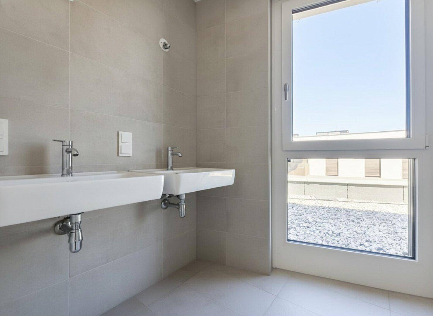 Modernes Badezimmer mit Doppelwaschbecken, hellen Fliesen und großem Fenster für Tageslicht.