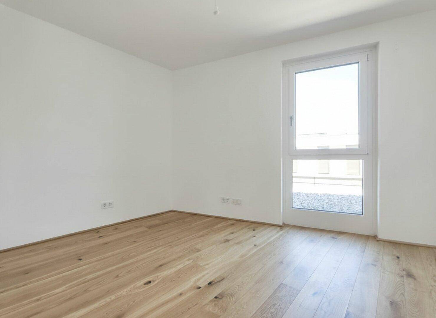 Helles Zimmer mit Holzdielenboden und großem Fenster, das viel Tageslicht hereinlässt.