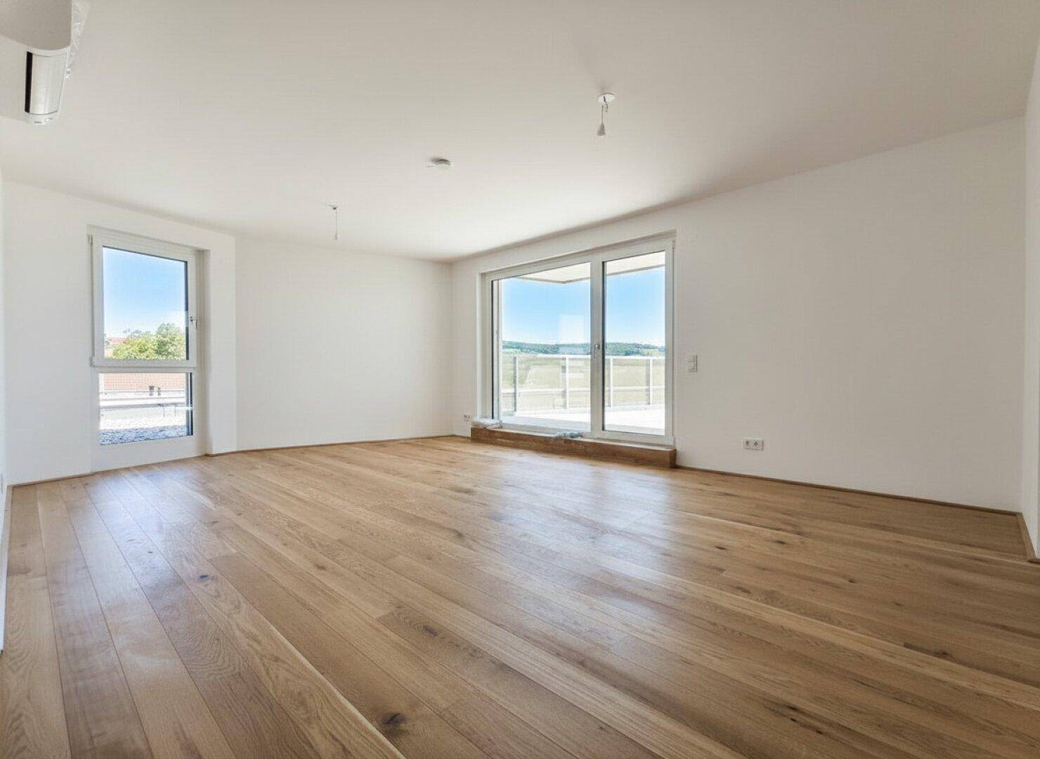 Lichtdurchflutetes Zimmer mit Holzdielen und großen Fenstern, die den Blick ins Grüne freigeben.