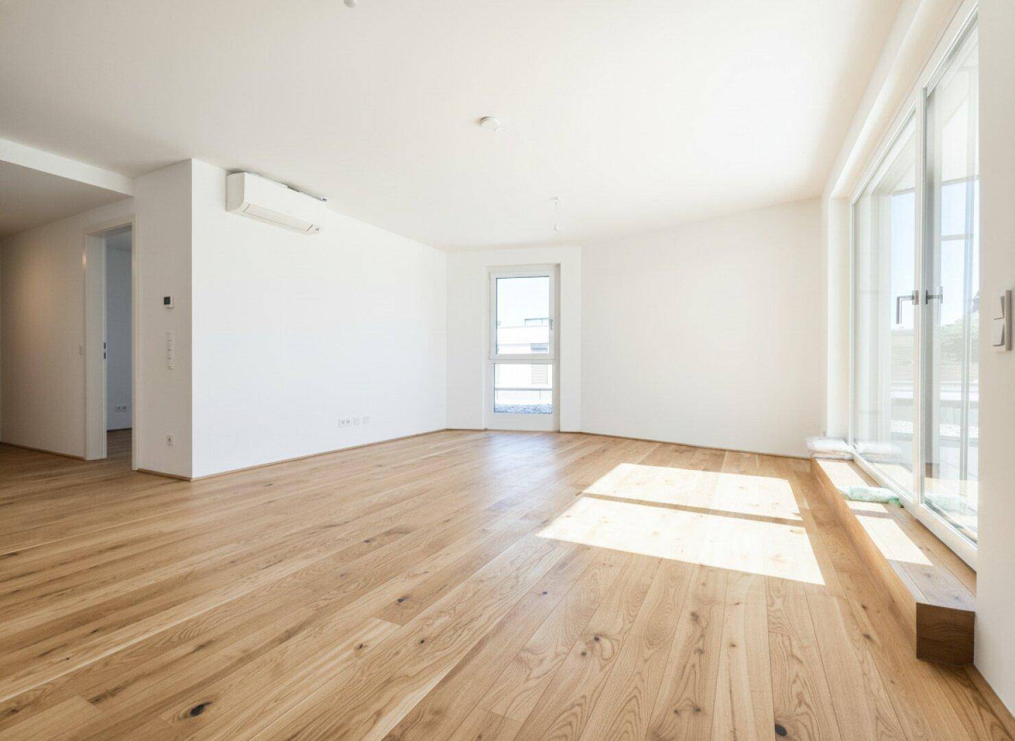 Helles, leeres Wohnzimmer mit breiten Holzdielen und Zugang zu einem Außenbereich.