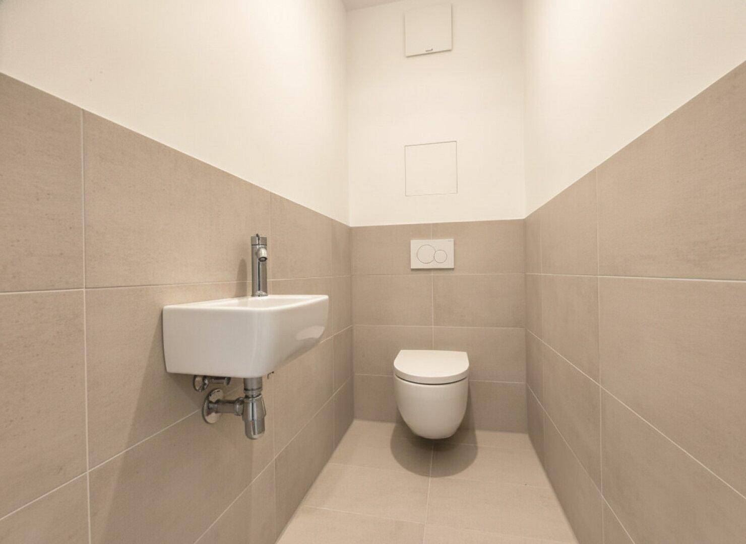 Gäste-WC mit beigefarbenen Fliesen, wandhängender Toilette und kleinem Handwaschbecken.