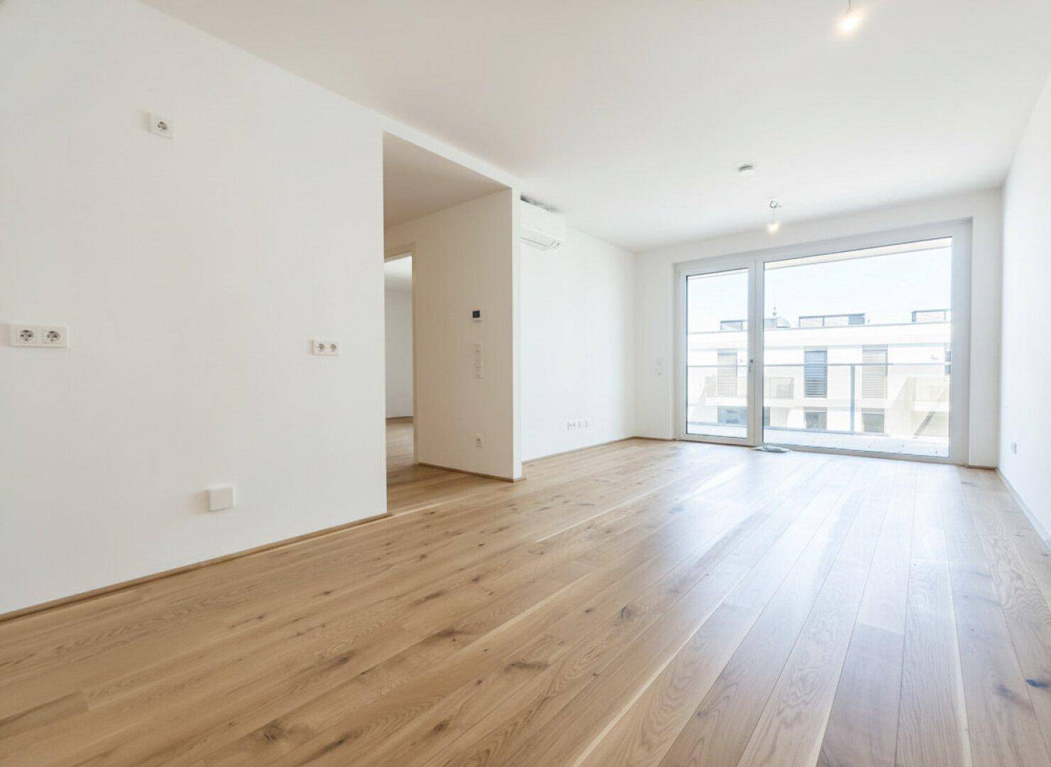 Helles, leeres Zimmer mit Holzboden, großen Fenstern und Zugang zu einem Balkon.