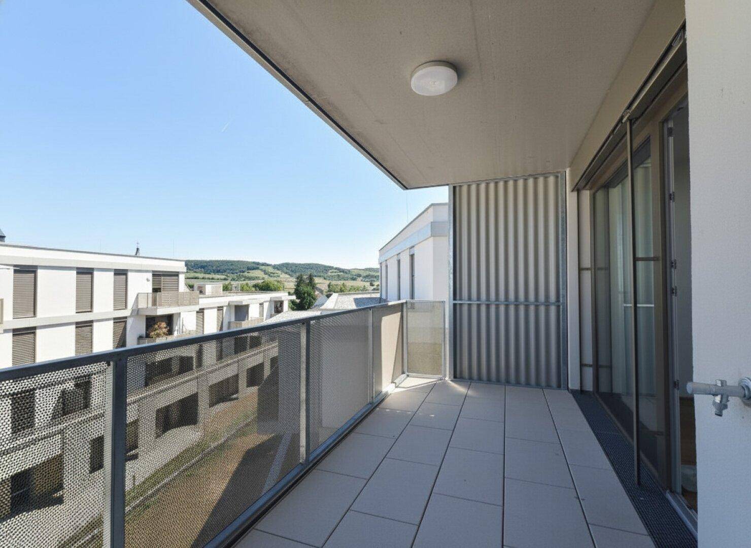 Geräumiger Balkon mit Fliesen und Blick auf die umliegenden Neubauten und grüne Hügel.