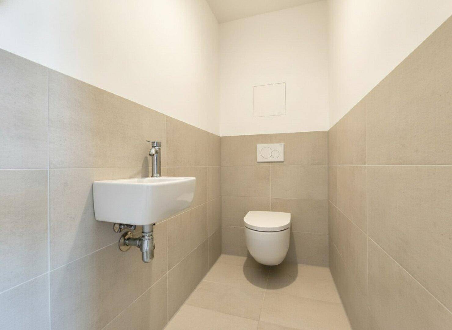 Modernes Gäste-WC mit hellgrauen Fliesen, wandhängendem WC und kleinem Handwaschbecken.